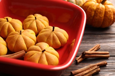 Photo of Baking tray with tasty pumpkin shaped buns and cinnamon sticks on wooden table, closeup