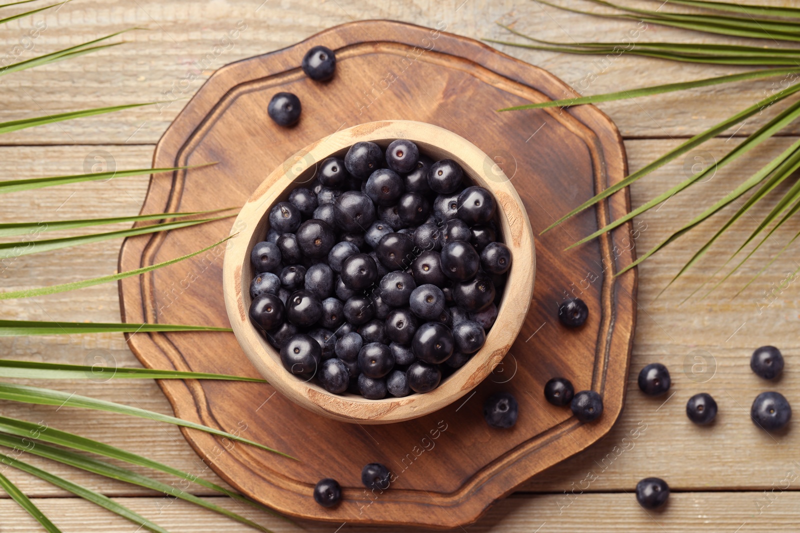 Photo of Ripe acai berries in bowl and palm leaves on wooden table, flat lay