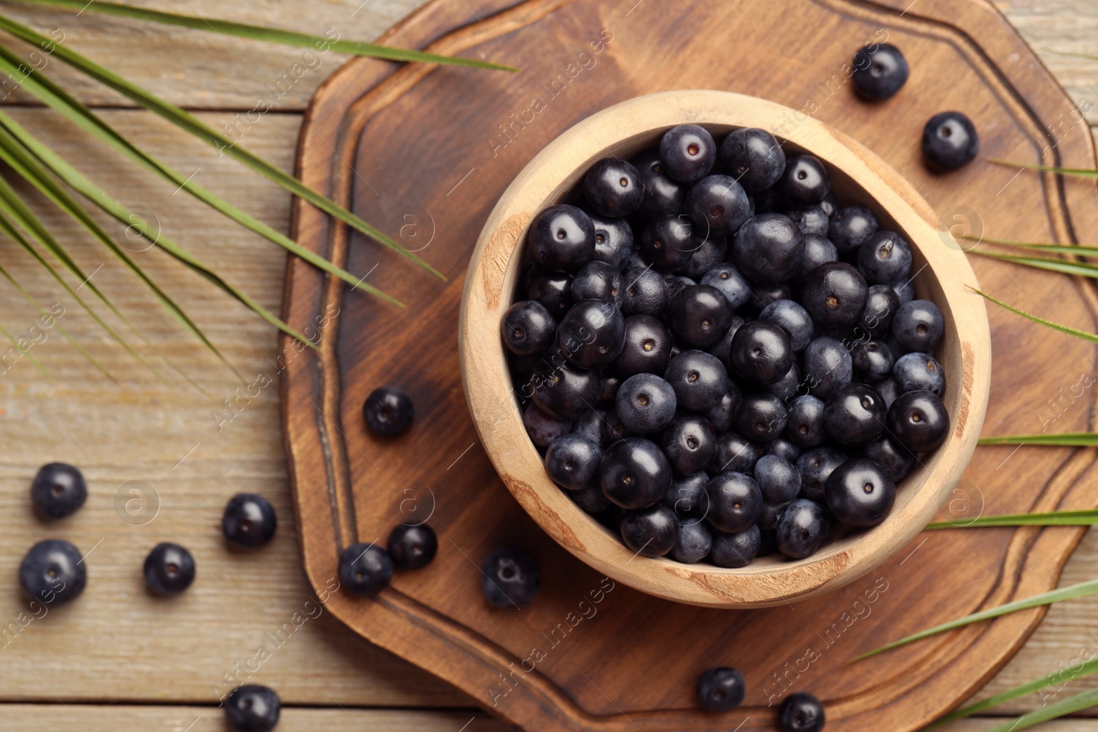 Photo of Ripe acai berries in bowl and palm leaves on wooden table, flat lay