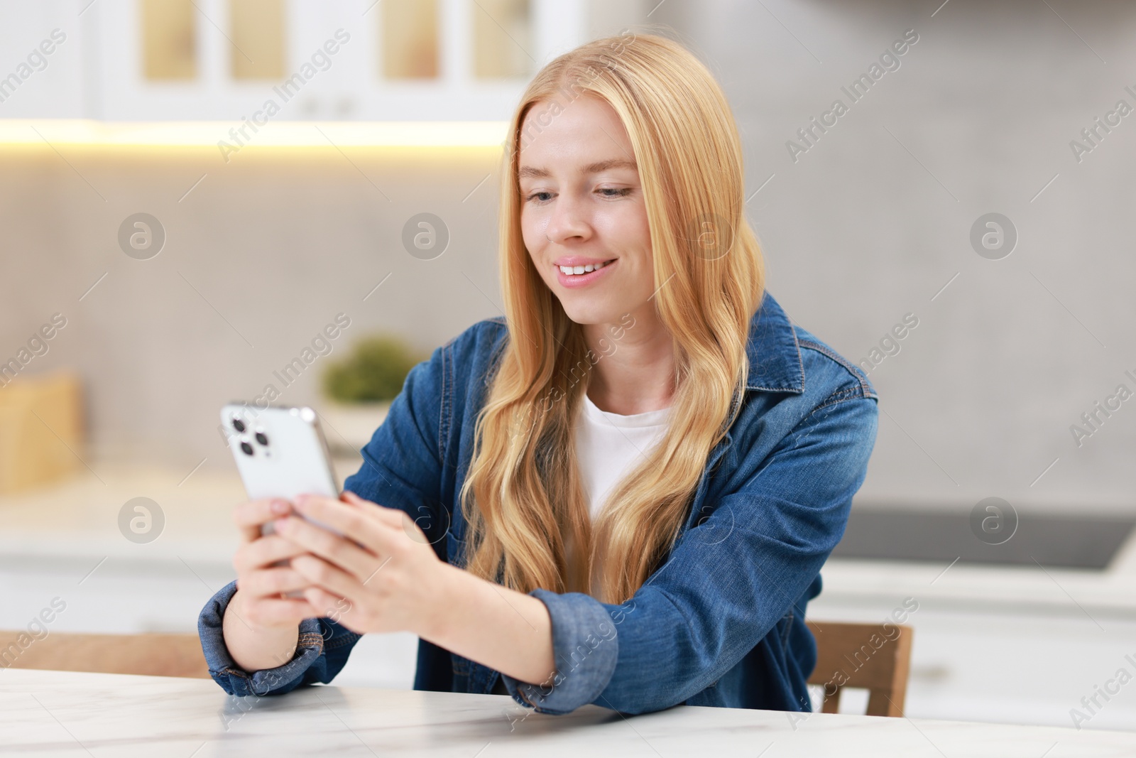 Photo of Smiling woman using smartphone at white table in kitchen