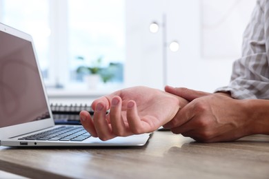 Photo of Man suffering from pain in wrist while working on laptop at table indoors, closeup. Carpal tunnel syndrome