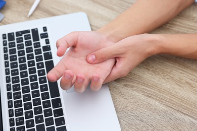 Photo of Man suffering from pain in wrist while working on laptop at wooden table, closeup. Carpal tunnel syndrome