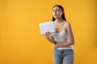 Woman holding calendar with marked menstrual cycle days and suffering from abdominal pain on orange background, space for text