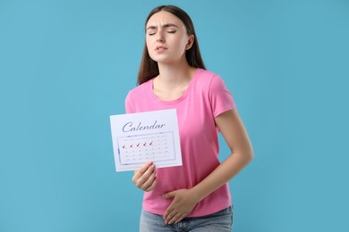 Photo of Woman holding calendar with marked menstrual cycle days and suffering from abdominal pain on light blue background