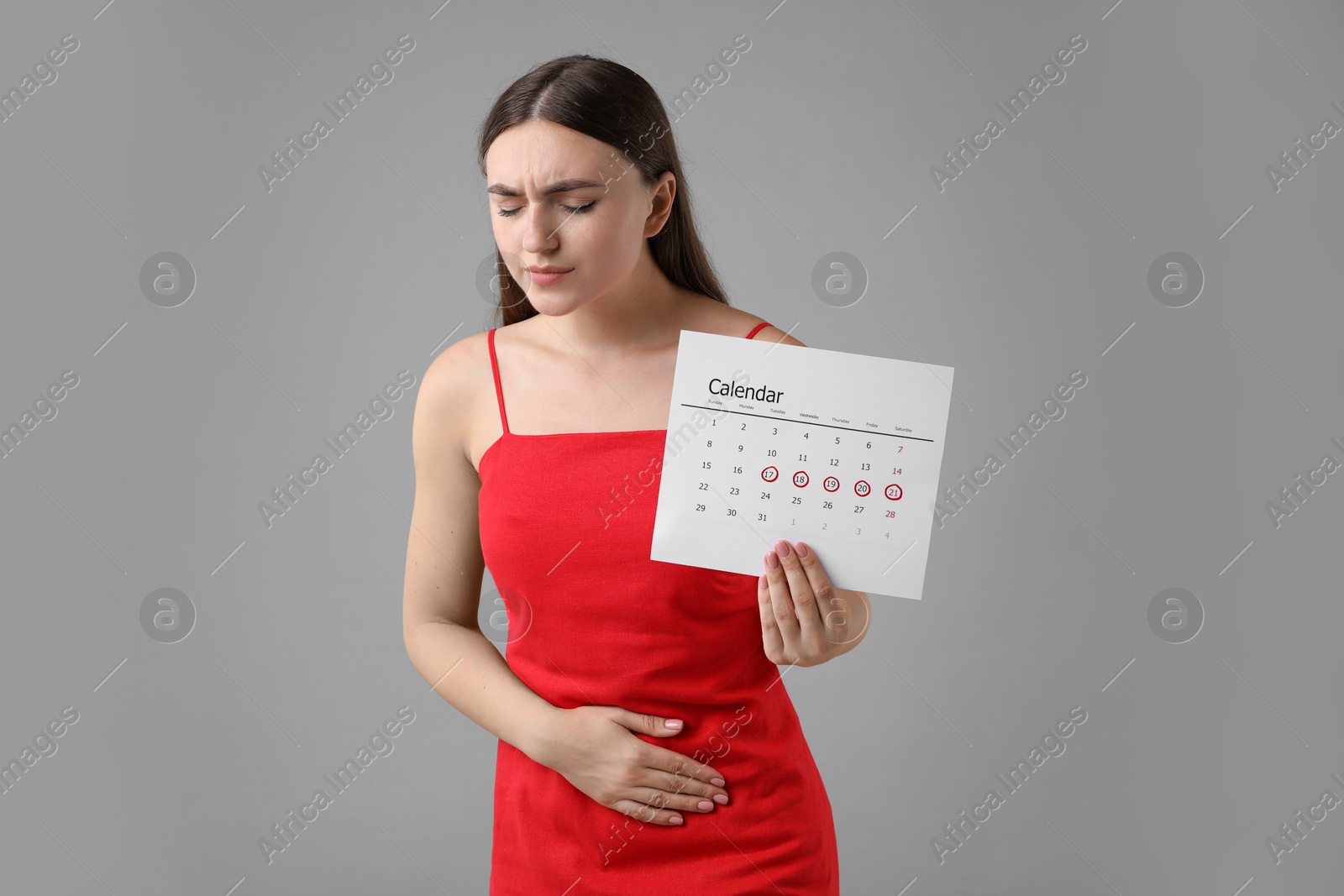 Photo of Woman holding calendar with marked menstrual cycle days and suffering from abdominal pain on grey background