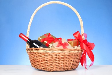 Photo of Wicker gift basket with wine on white marble table against light blue background