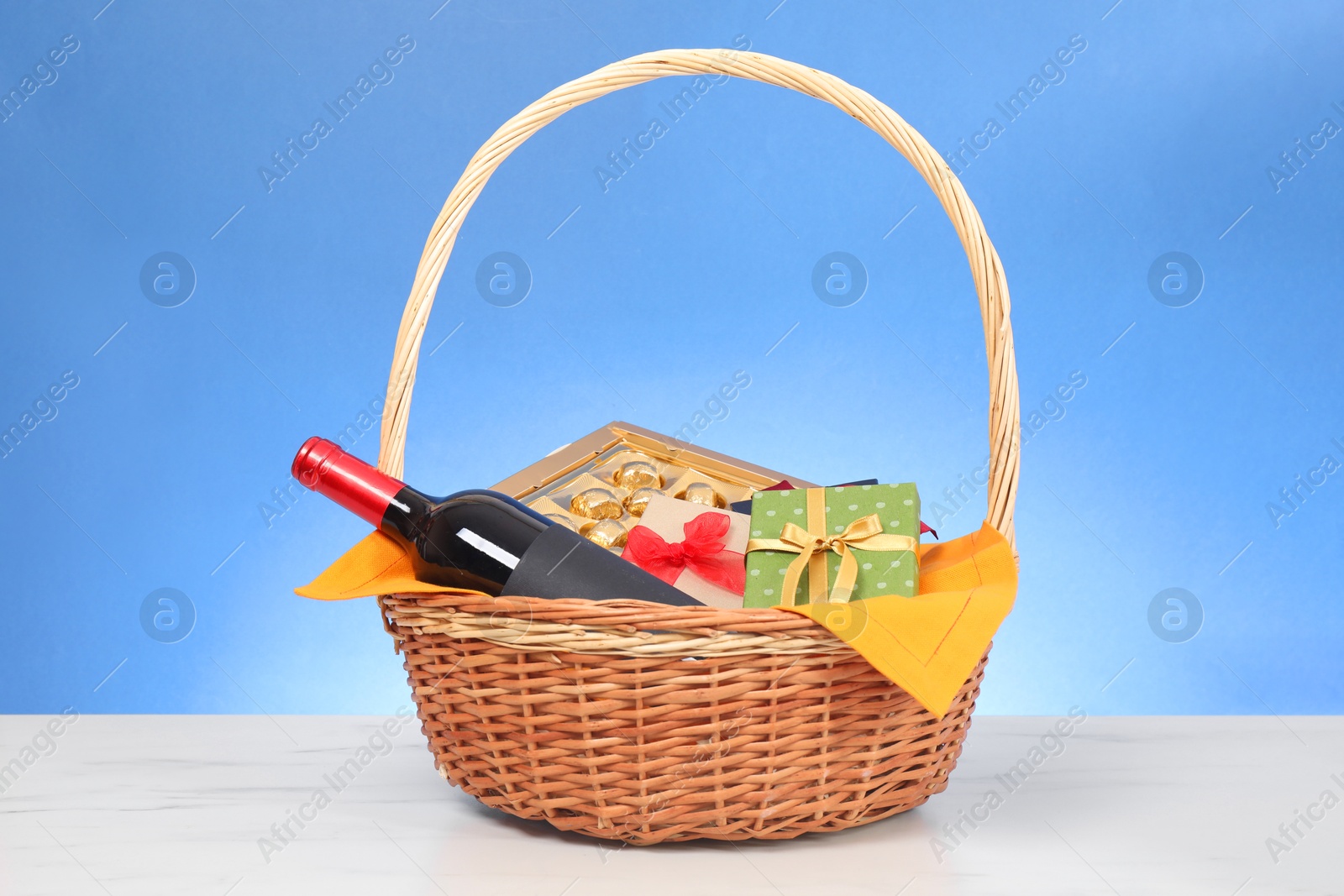 Photo of Wicker gift basket with wine on white marble table against light blue background