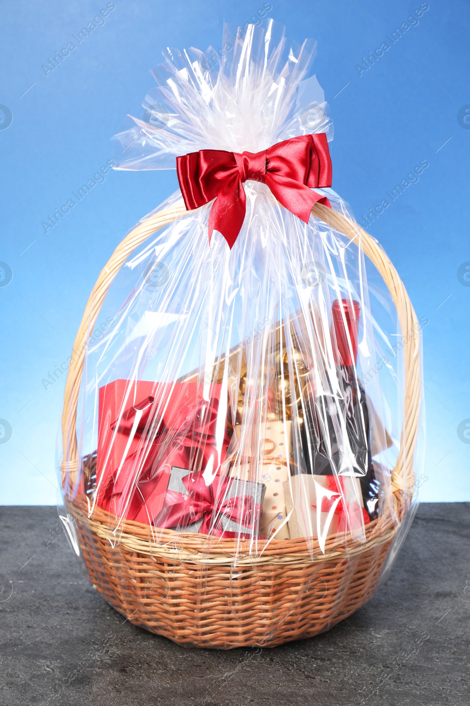 Photo of Wicker gift basket with wine on grey textured table against light blue background