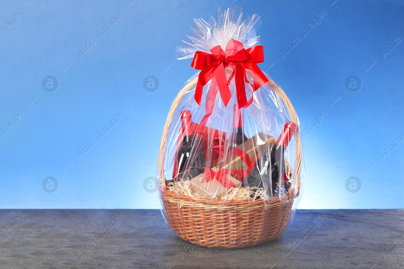 Photo of Wicker gift basket with wine on grey textured table against light blue background