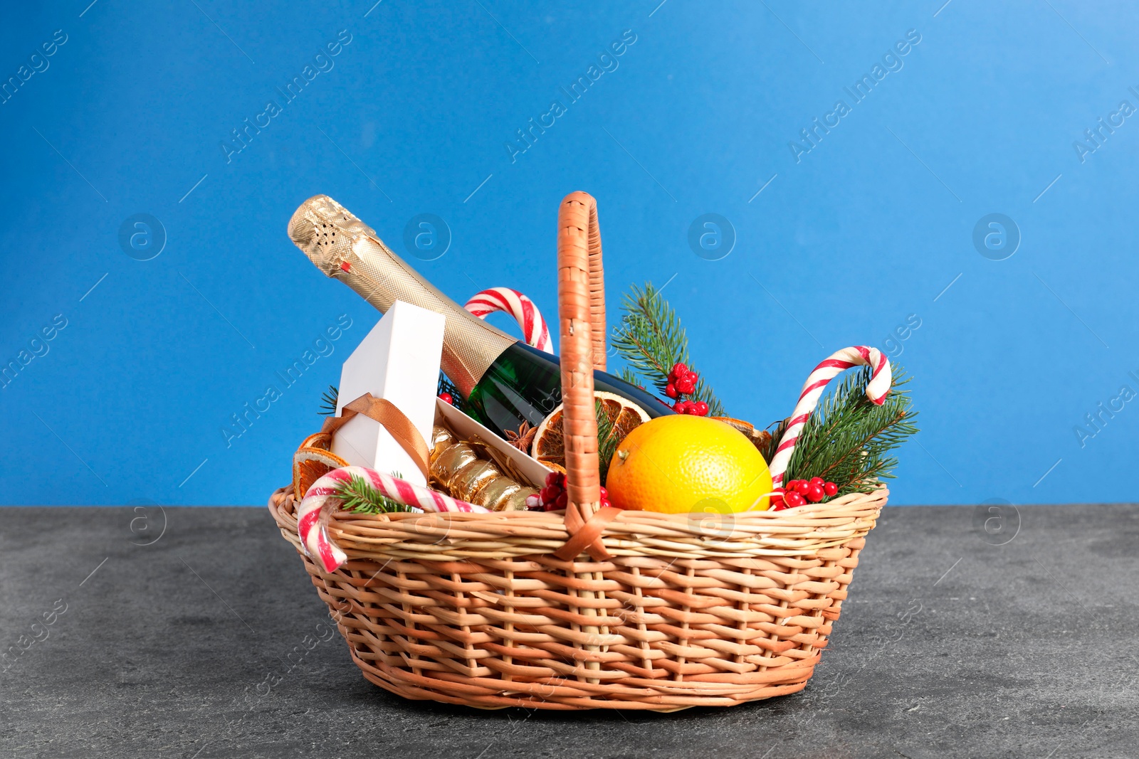 Photo of Wicker gift basket with sparkling wine and Christmas decor on grey textured table against light blue background