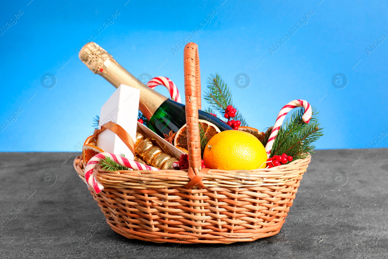 Photo of Wicker gift basket with sparkling wine and Christmas decor on grey textured table against light blue background