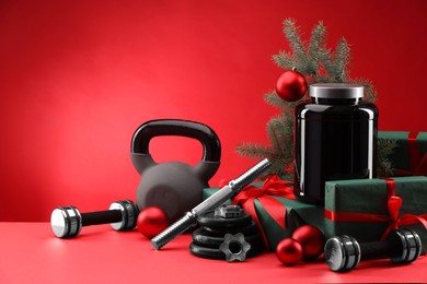 Photo of Dumbbells, kettlebell, container with supplement and Christmas decor on red background. Space for text