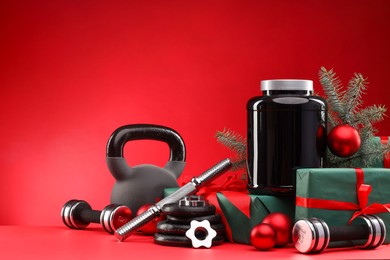 Photo of Dumbbells, kettlebell, container with supplement and Christmas decor on red background. Space for text