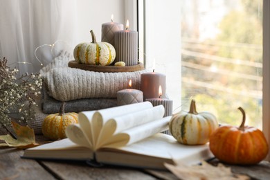 Photo of Open book, stack of sweaters, burning candles and pumpkins on window sill. Autumn atmosphere