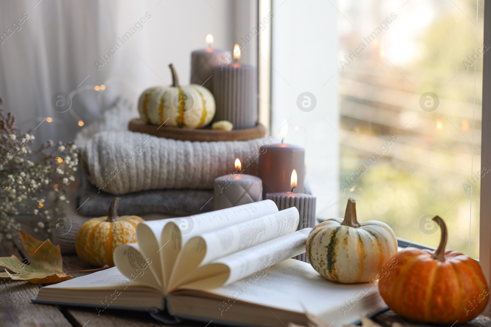 Photo of Open book, stack of sweaters, burning candles and pumpkins on window sill. Autumn atmosphere