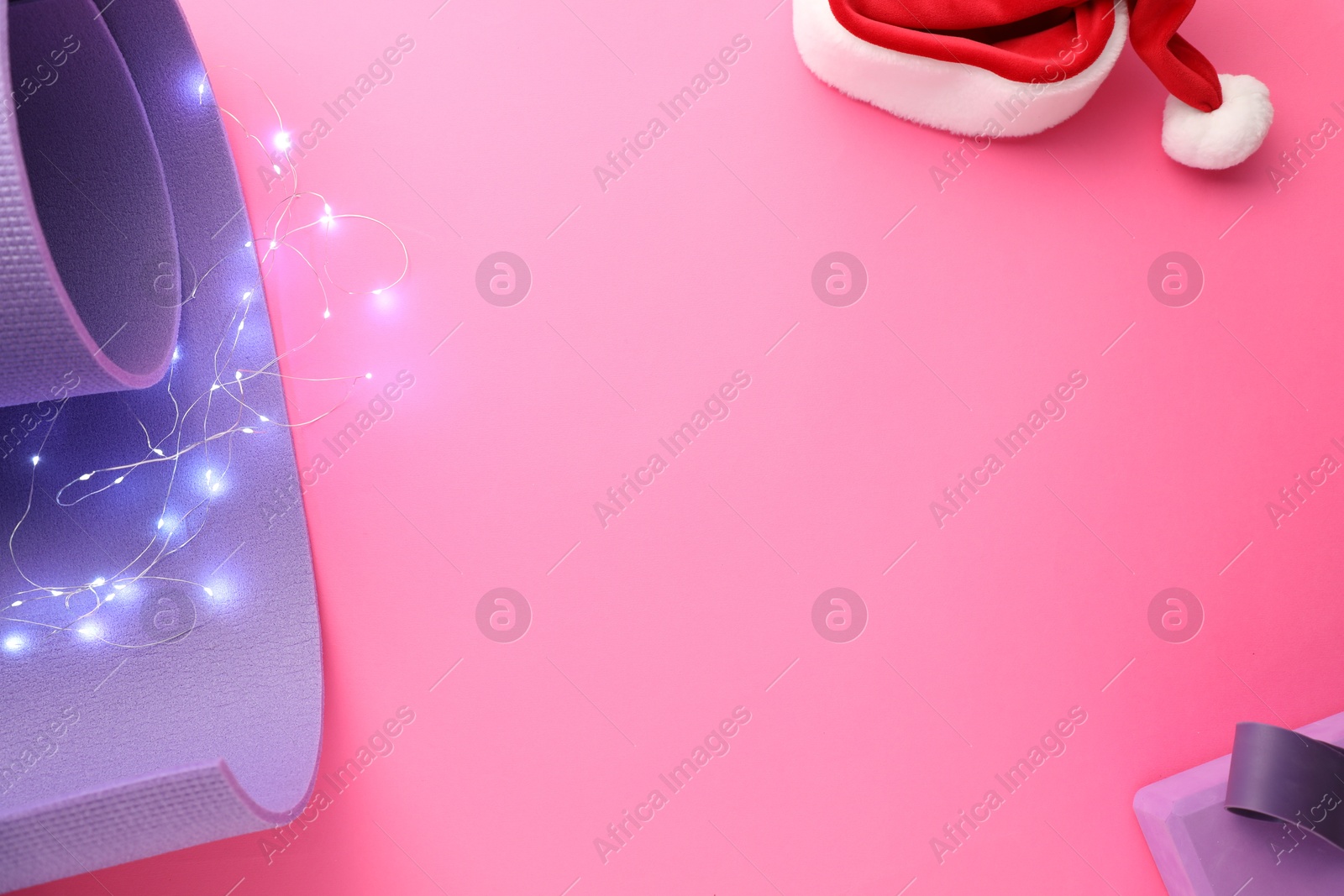 Photo of Yoga mat, block, elastic band, Santa hat and Christmas lights on pink background, flat lay. Space for text