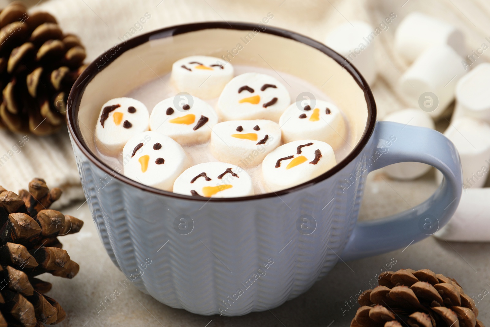 Photo of Marshmallows with snowmen faces in cup of hot drink on light table, closeup