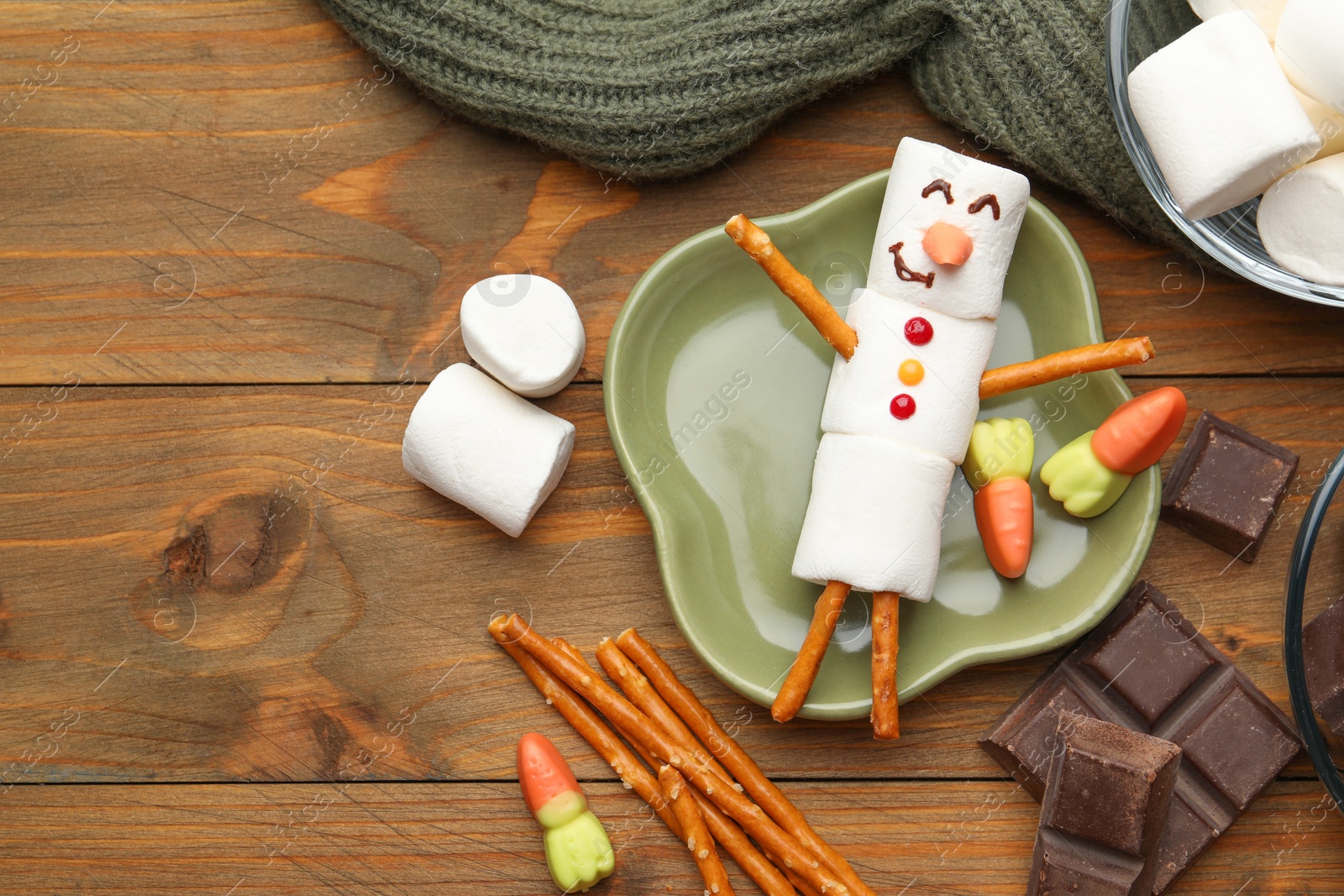 Photo of Funny snowman made with marshmallows, candies, chocolate and salty sticks on wooden table, flat lay