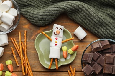 Photo of Funny snowman made with marshmallows, candies, chocolate and salty sticks on wooden table, flat lay