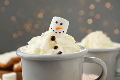 Photo of Funny snowman made with marshmallow and whipped cream in cup of hot drink against Christmas lights, closeup