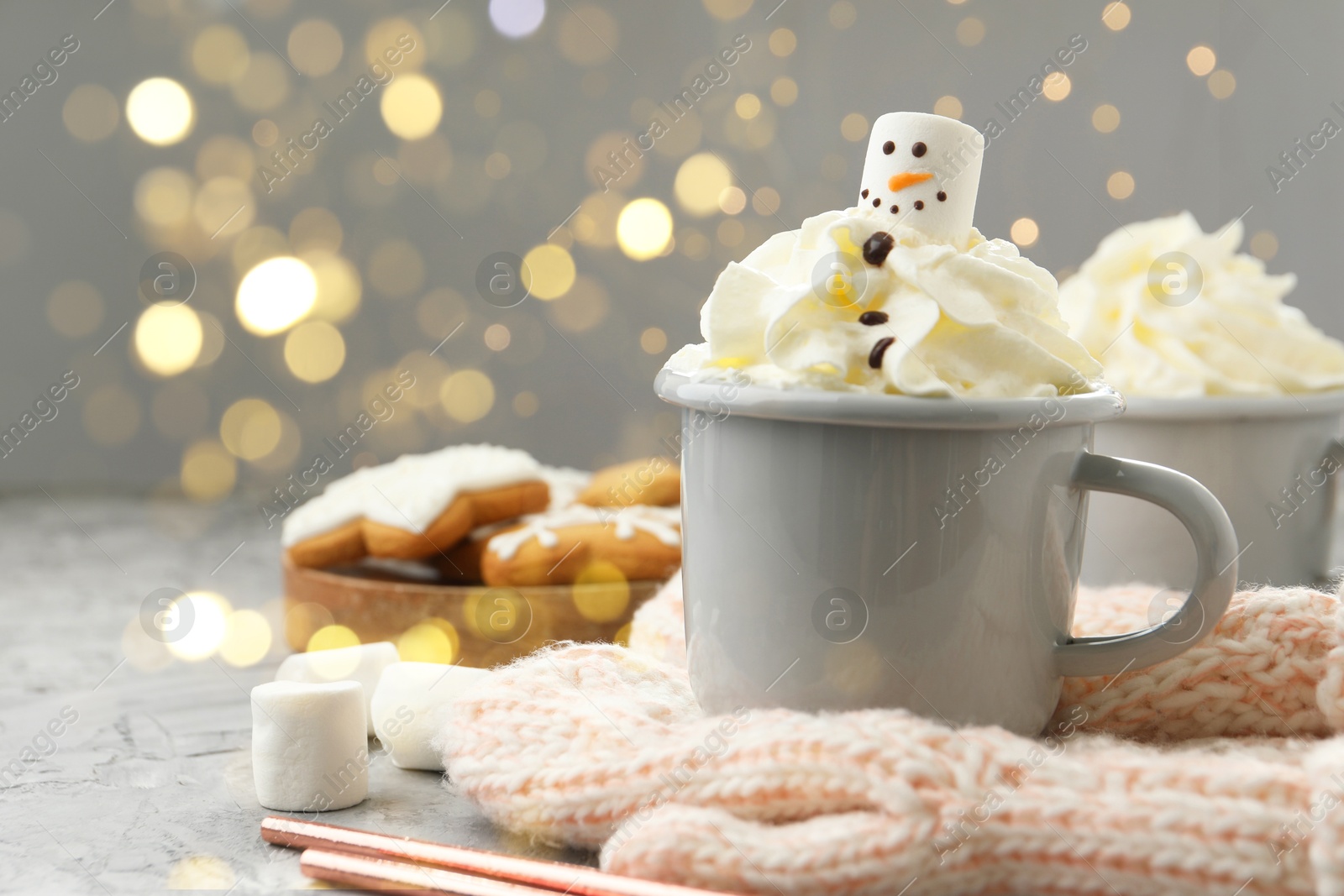 Photo of Funny snowman made with marshmallow and whipped cream in cup of hot drink served on grey background against Christmas lights, closeup. Space for text