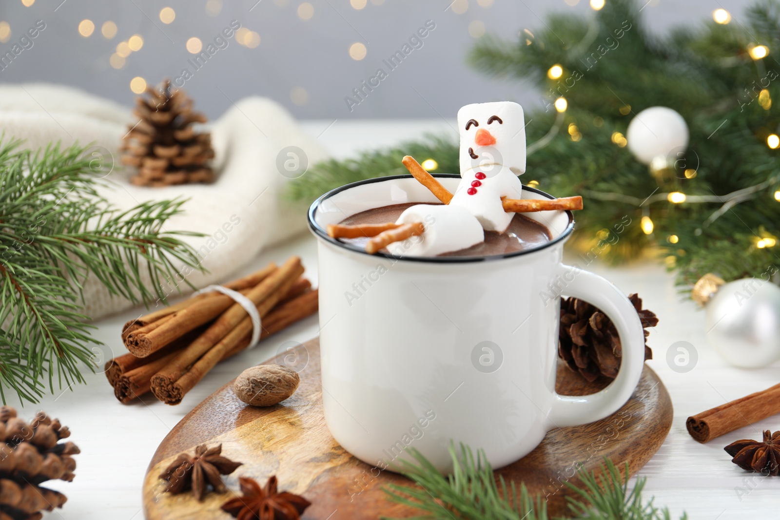 Photo of Funny snowman made with marshmallows in cup of hot chocolate and winter decor on white table against Christmas lights