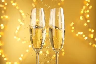 Photo of Glasses of sparkling wine and confetti against golden background with blurred lights, closeup. Christmas decor