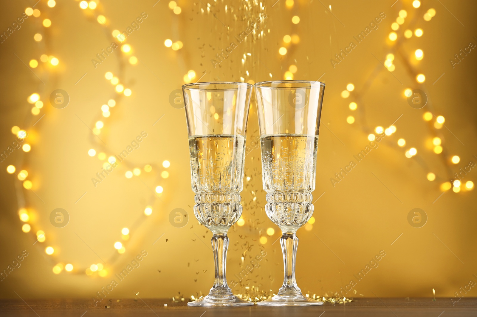 Photo of Glasses of sparkling wine and confetti against golden background with blurred lights. Christmas decor