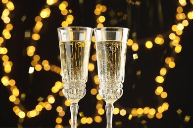 Photo of Glasses of sparkling wine and confetti against dark background with blurred lights, closeup. Christmas decor