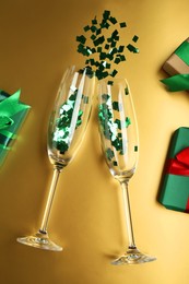 Photo of Wine glasses with confetti and gift boxes on golden background, flat lay. Christmas decor