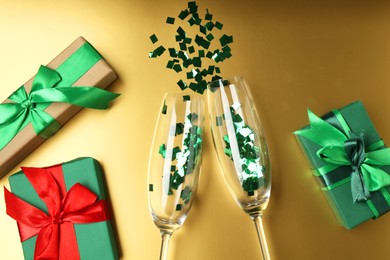 Photo of Wine glasses with confetti and gift boxes on golden background, flat lay. Christmas decor