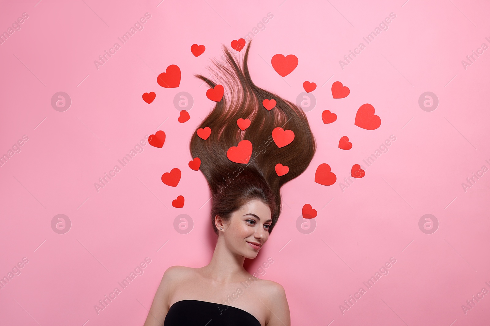 Photo of Beautiful young woman with long hair and red paper hearts on pink background, top view