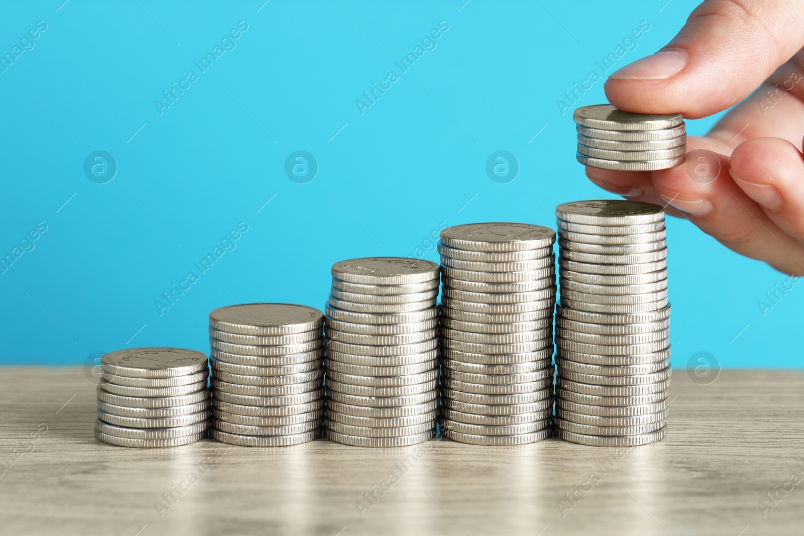 Photo of Woman putting coins at wooden table against light blue background, closeup. Salary concept