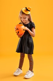 Photo of Cute girl with pumpkin bucket dressed like witch on yellow background. Halloween celebration