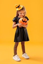 Photo of Cute girl with pumpkin bucket dressed like witch on yellow background. Halloween celebration