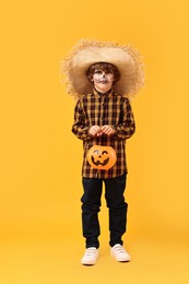 Funny boy with pumpkin bucket dressed like scarecrow on yellow background. Halloween celebration