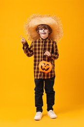 Photo of Funny boy with pumpkin bucket dressed like scarecrow pointing at something on yellow background. Halloween celebration