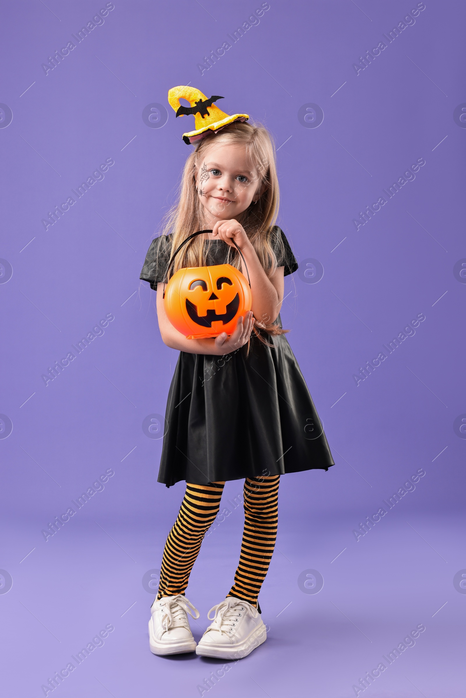 Photo of Cute girl with pumpkin bucket dressed like witch on violet background. Halloween celebration