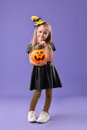 Photo of Cute girl with pumpkin bucket dressed like witch on violet background. Halloween celebration