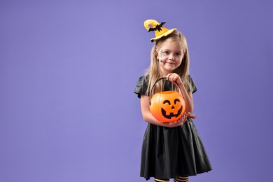 Photo of Cute girl with pumpkin bucket dressed like witch on violet background, space for text. Halloween celebration