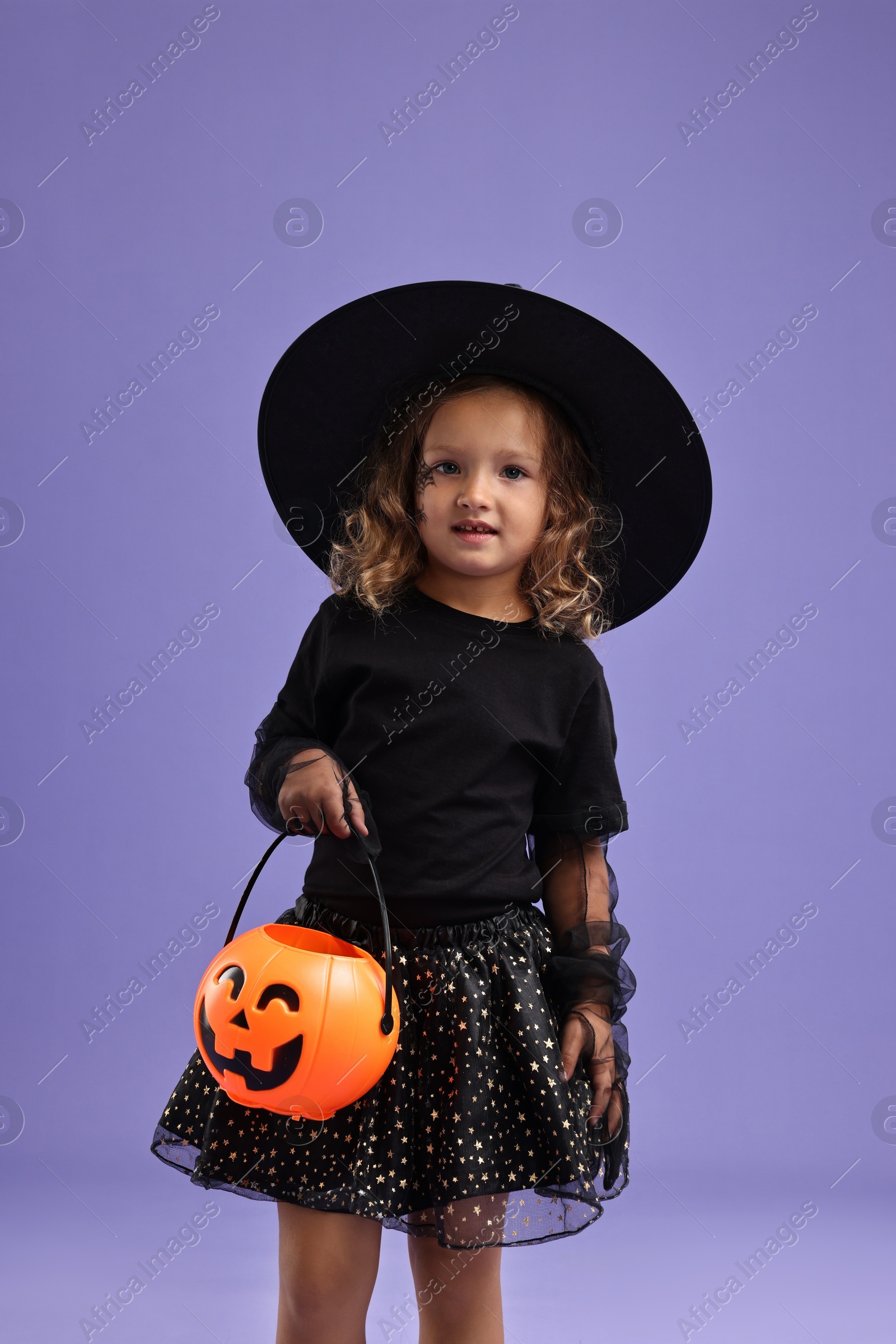 Photo of Cute girl with pumpkin bucket dressed like witch on violet background. Halloween costume