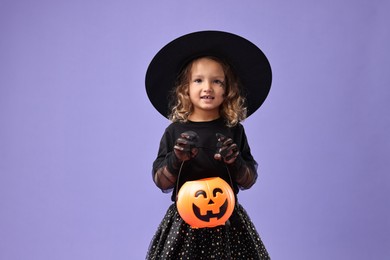 Photo of Cute girl with pumpkin bucket dressed like witch on violet background. Halloween costume