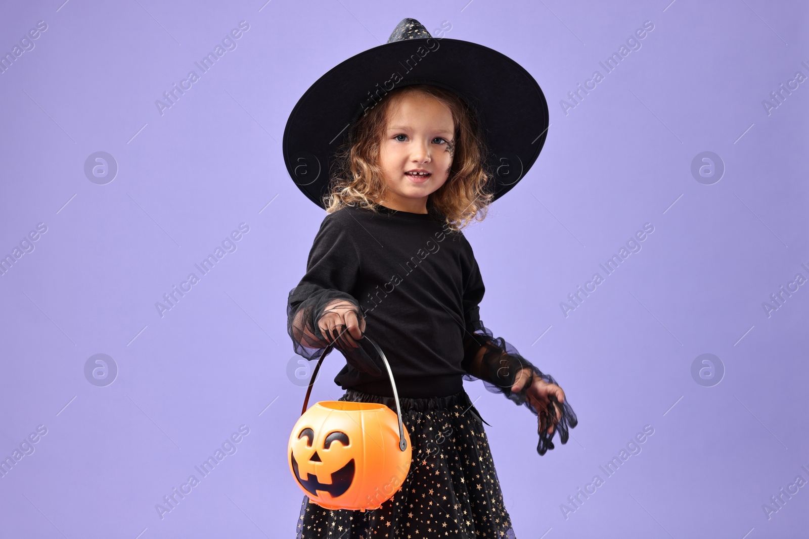 Photo of Cute girl with pumpkin bucket dressed like witch on violet background. Halloween costume