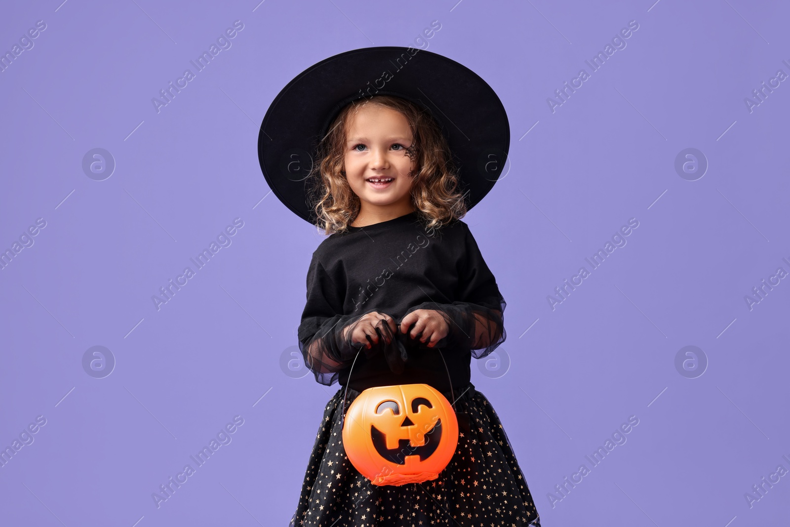 Photo of Cute girl with pumpkin bucket dressed like witch on violet background. Halloween costume