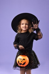 Cute girl with pumpkin bucket dressed like witch on violet background. Halloween costume