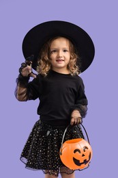 Photo of Cute girl with pumpkin bucket dressed like witch on violet background. Halloween costume