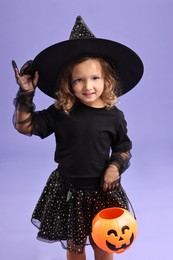 Photo of Cute girl with pumpkin bucket dressed like witch on violet background. Halloween costume