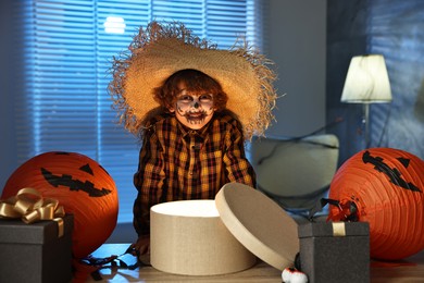 Photo of Cute boy dressed like scarecrow with festive decor and gift boxes indoors at night. Halloween celebration