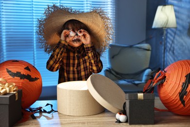 Photo of Cute boy dressed like scarecrow with decorative eyeballs, festive decor and gift boxes indoors at night. Halloween celebration