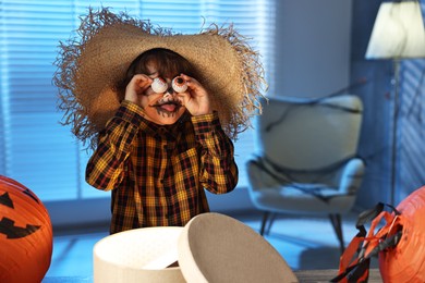 Photo of Boy dressed like scarecrow with decorative eyeballs, festive decor and gift box indoors at night. Halloween celebration